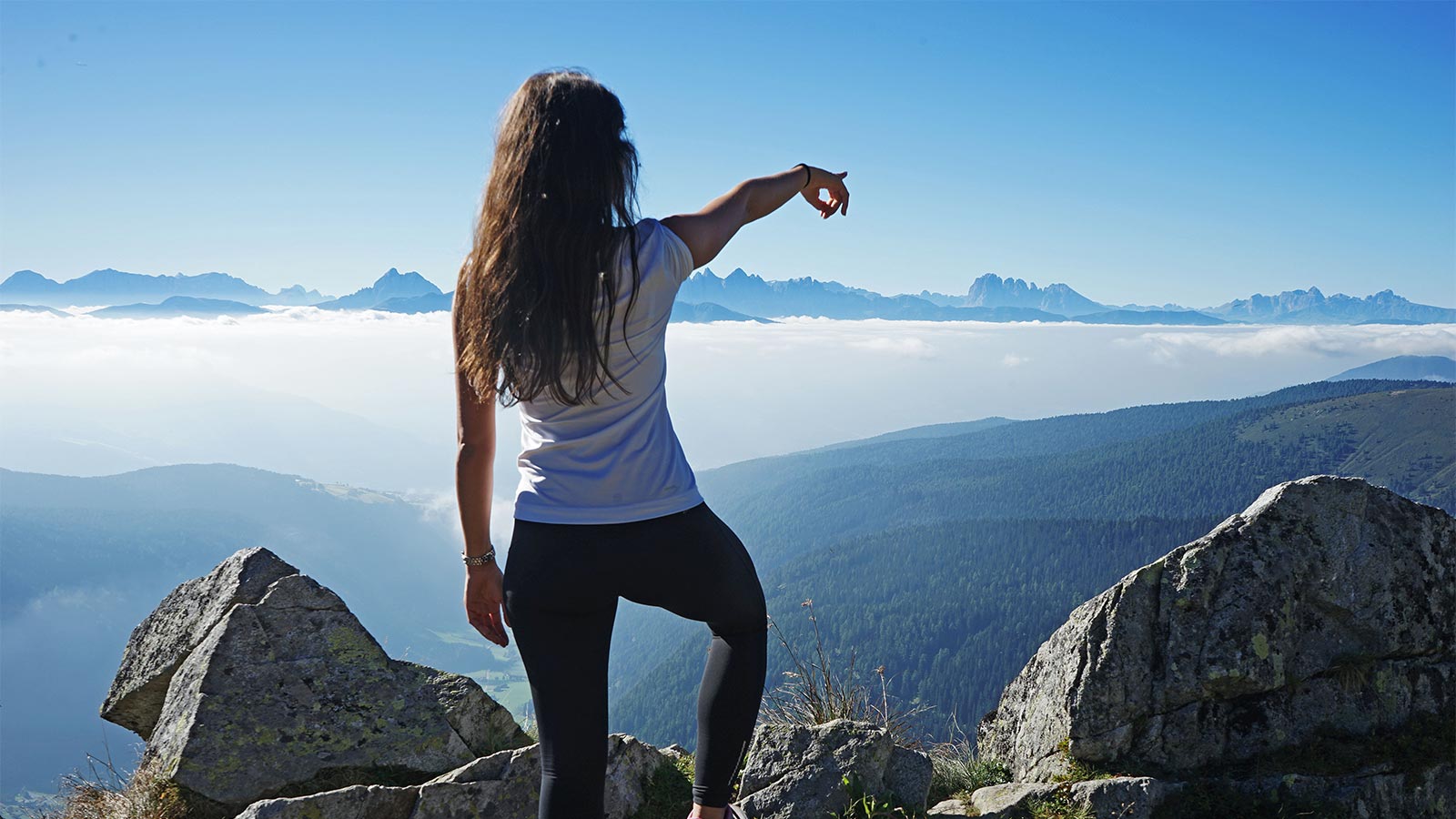 una giovane signora in cima alla montagna punta il dito verso l'orizzonte con altre cime nello sfondo